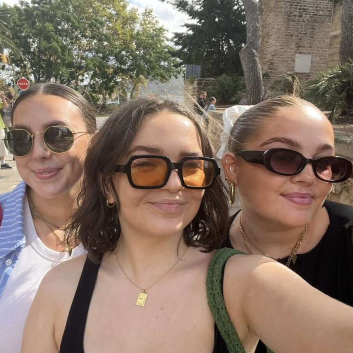 Three women taking a selfie outdoors The central woman is wearing oversized square sunglasses with orange-tinted lenses and a black top, flanked by two other women wearing sunglasses.