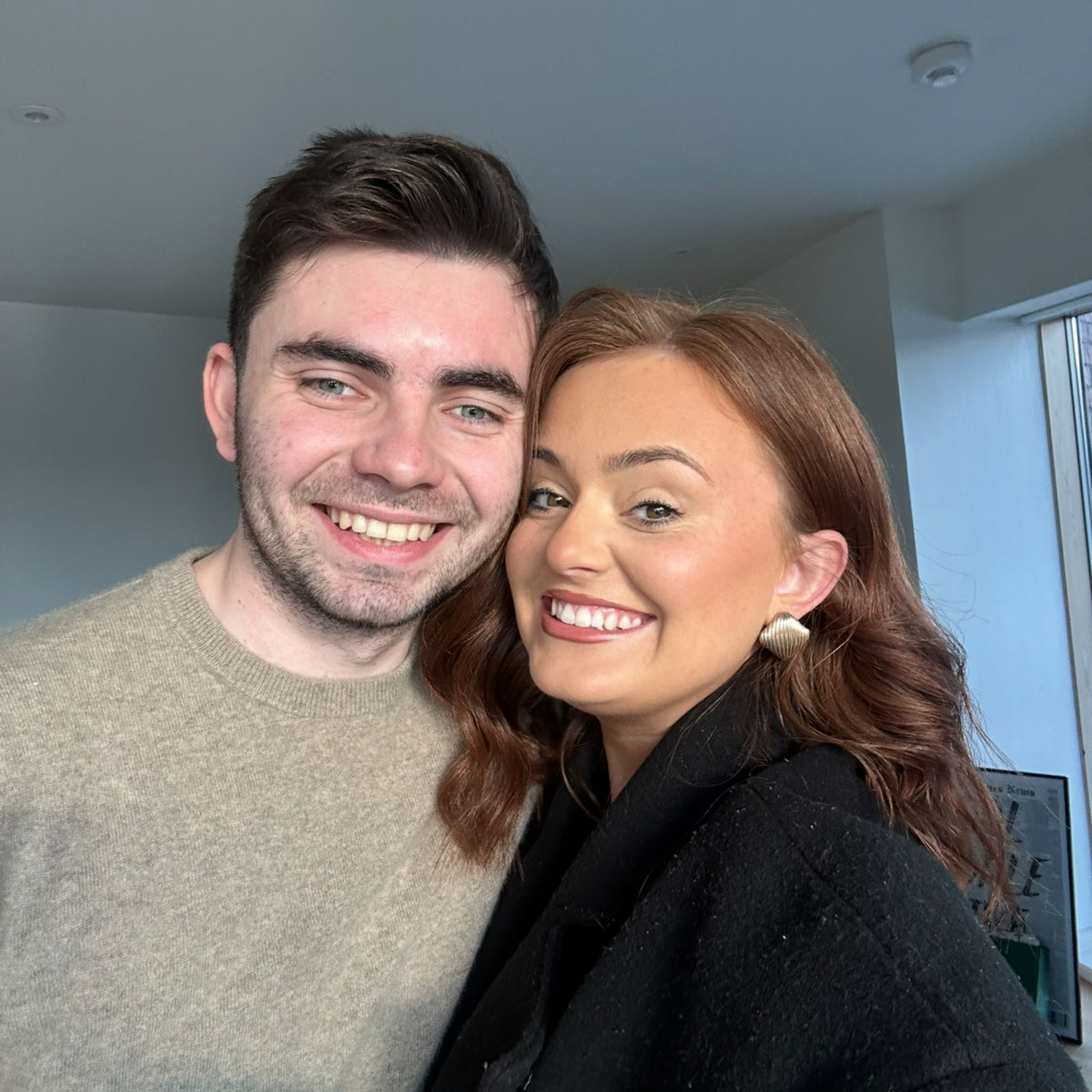 A smiling man and woman posing closely together indoors. The man wears a light beige knit sweater, and the woman wears a black coat and a small gold earrings.
