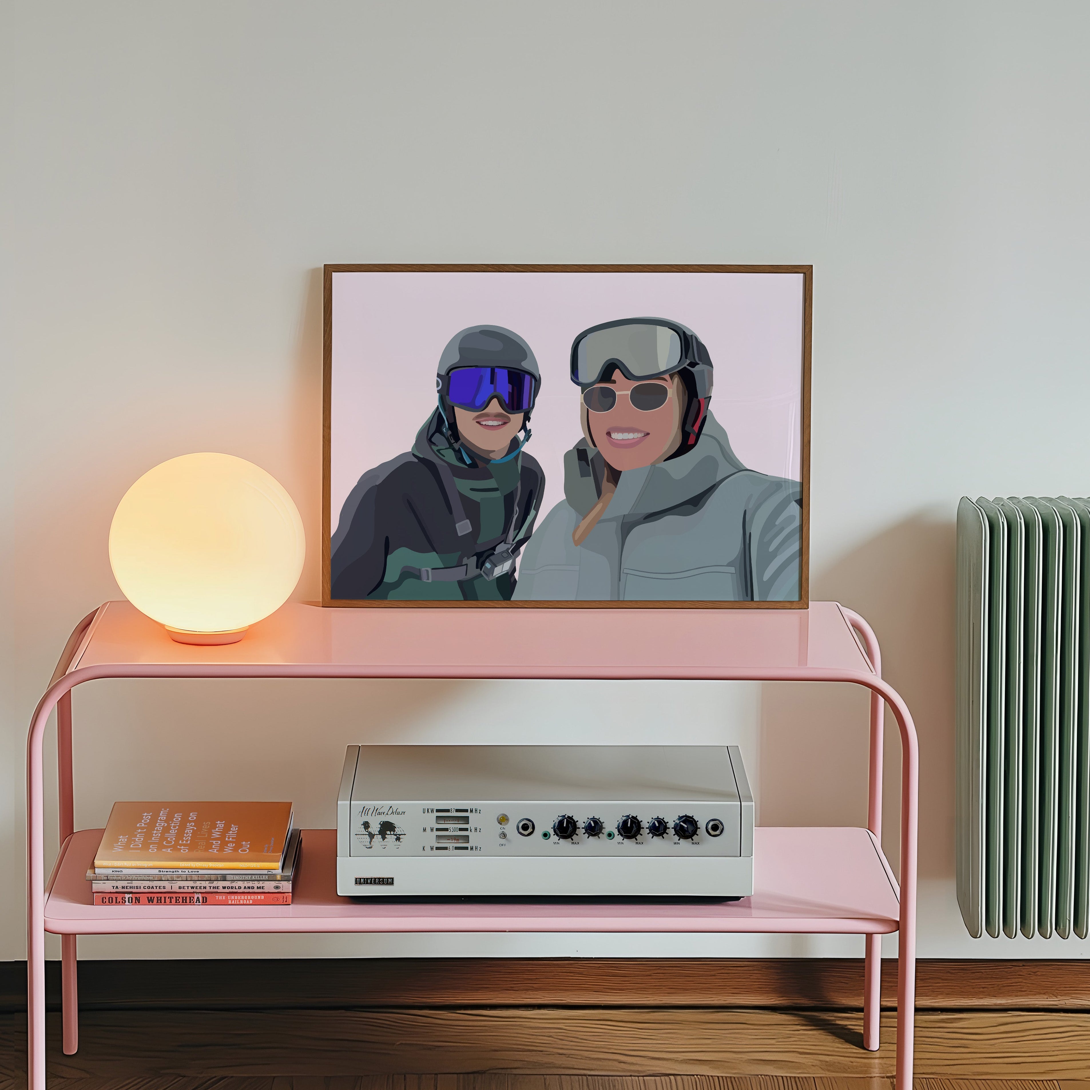 Pink console table with a lamp, books, and a vintage radio against a white wall with a framed Faceless Portrait of two people in skiing gear.