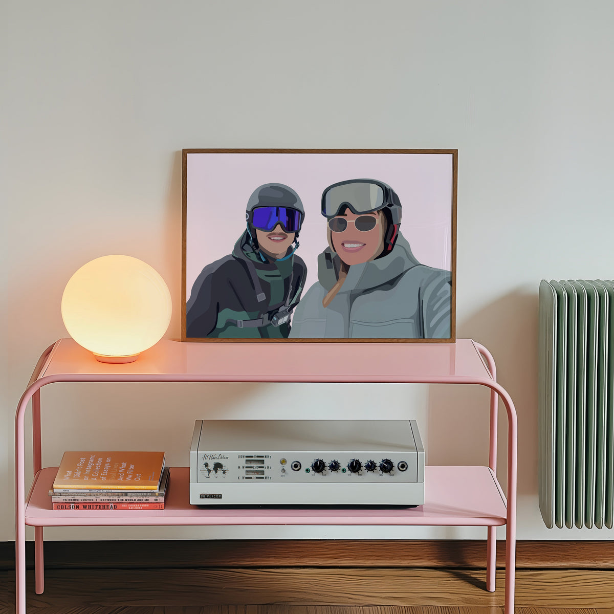 Pink console table with a lamp, books, and a vintage radio against a white wall with a framed Faceless Portrait of two people in skiing gear.