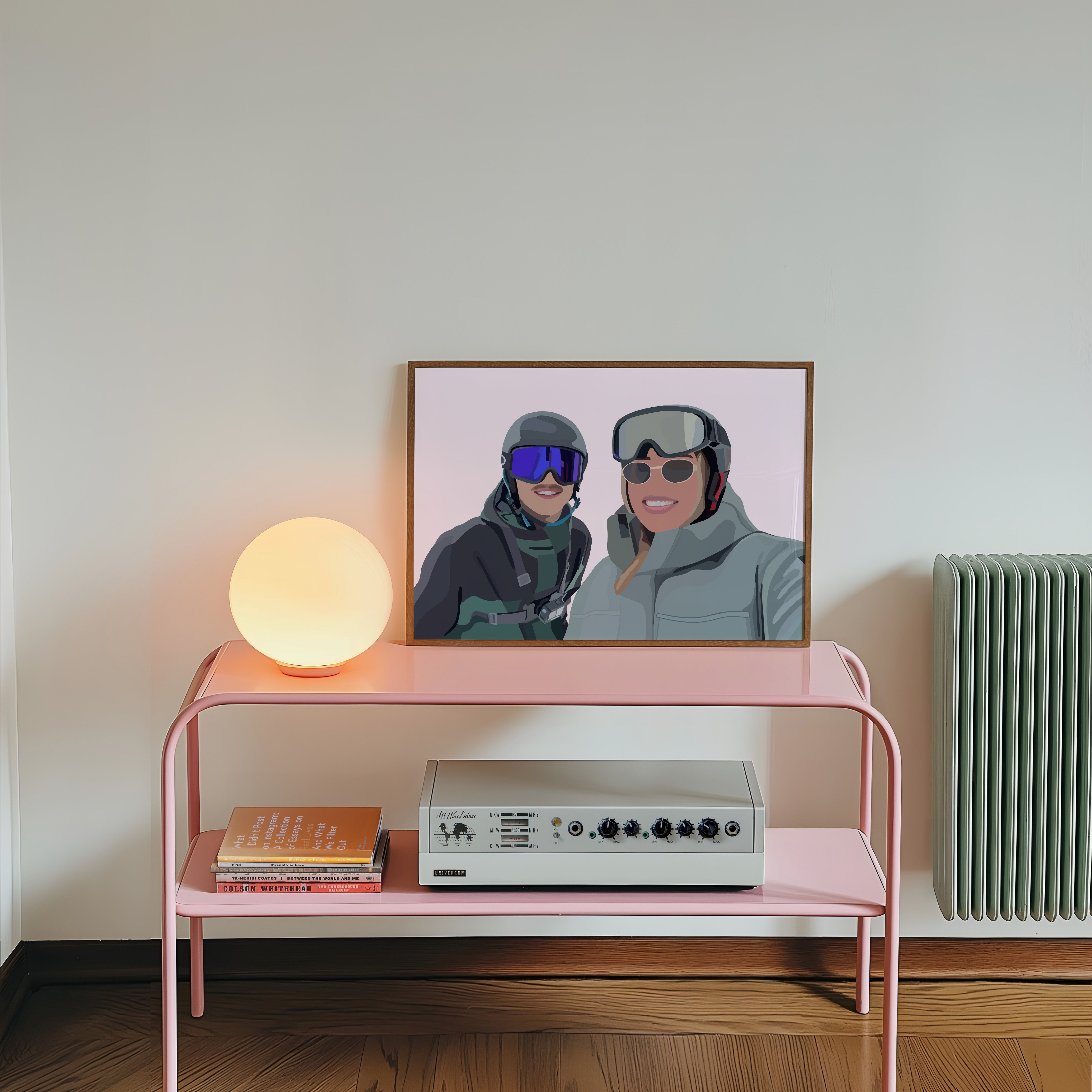 Pink console table with a lamp, books, and a vintage radio against a white wall with a framed Faceless Portrait of two people in skiing gear.