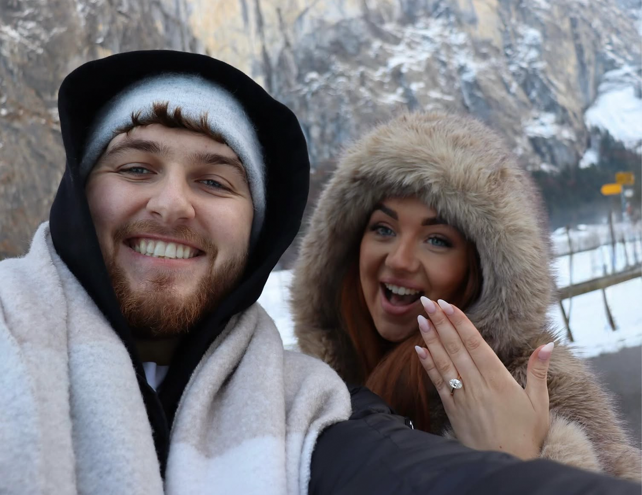 A smiling man and woman taking a selfie outdoors in winter clothing, with snow-covered mountains in the background. The woman is showing an engagement ring on her hand.