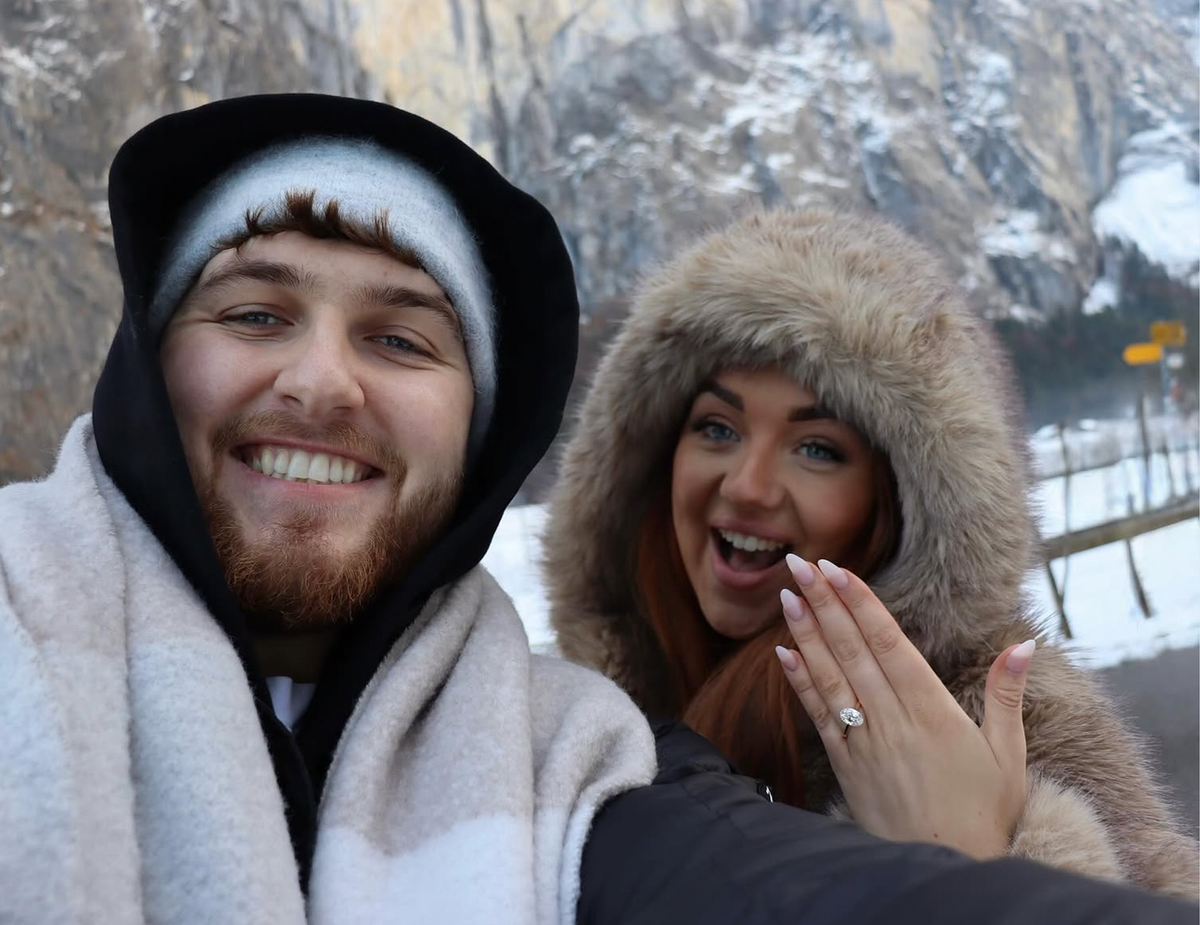 A smiling man and woman taking a selfie outdoors in winter clothing, with snow-covered mountains in the background. The woman is showing an engagement ring on her hand.