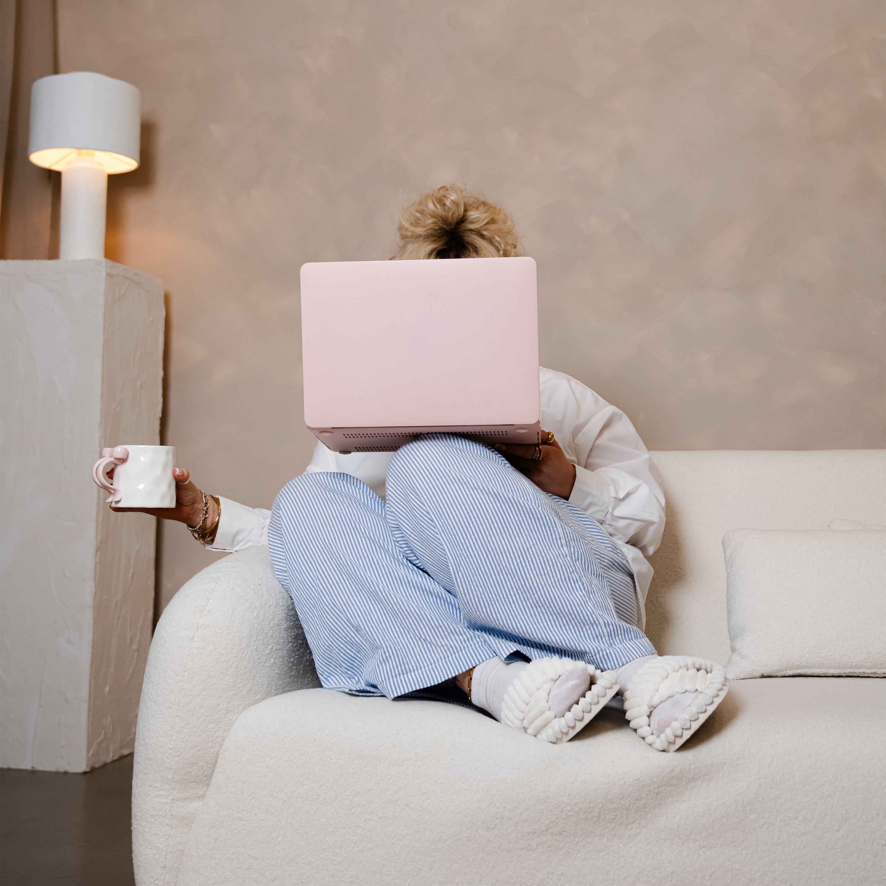 Person sitting on a sofa with a pink laptop over their face, holding a mug.