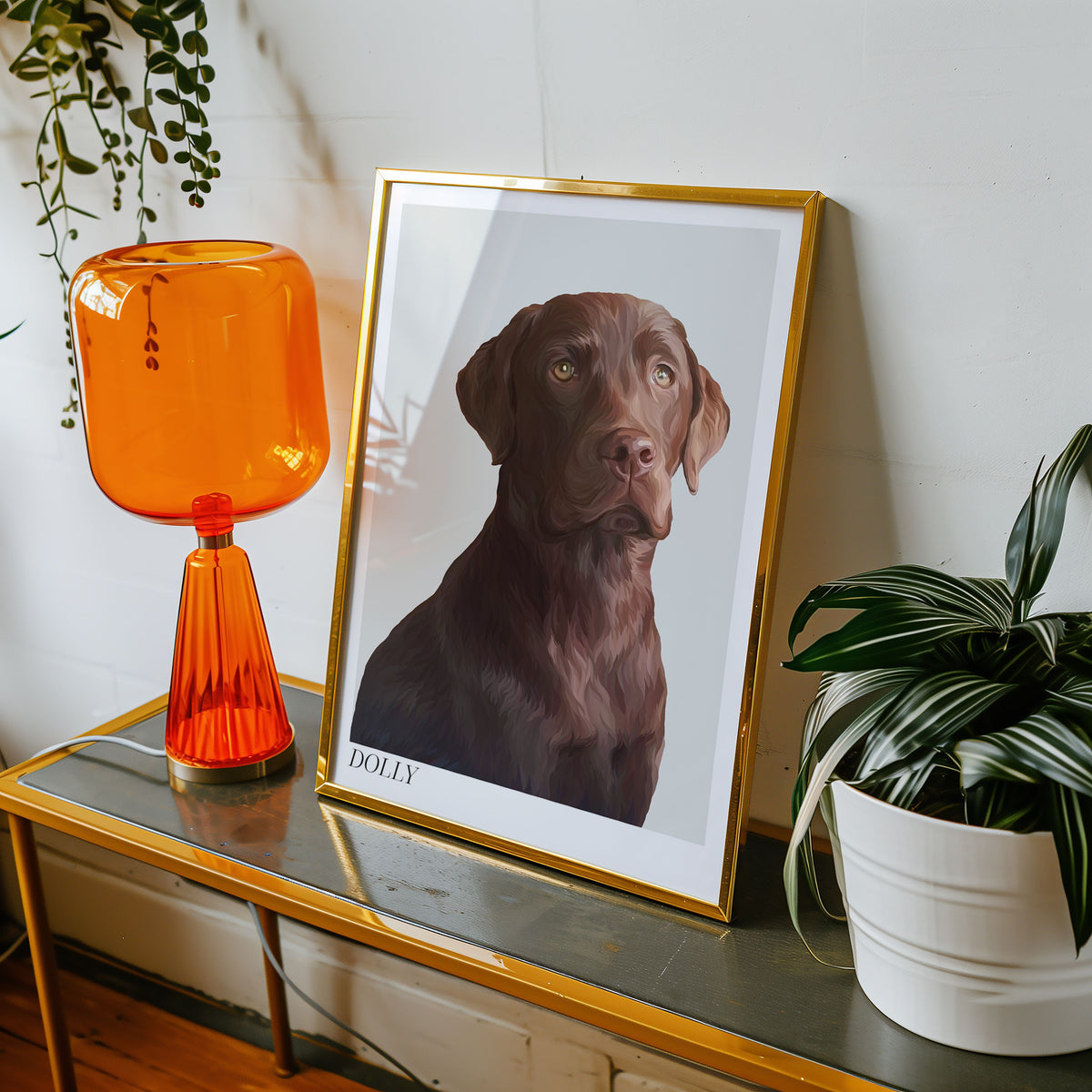 A framed Bordered Detailed Pet Portrait of a brown Labrador displayed on a table with an orange lamp and a plant in the background.