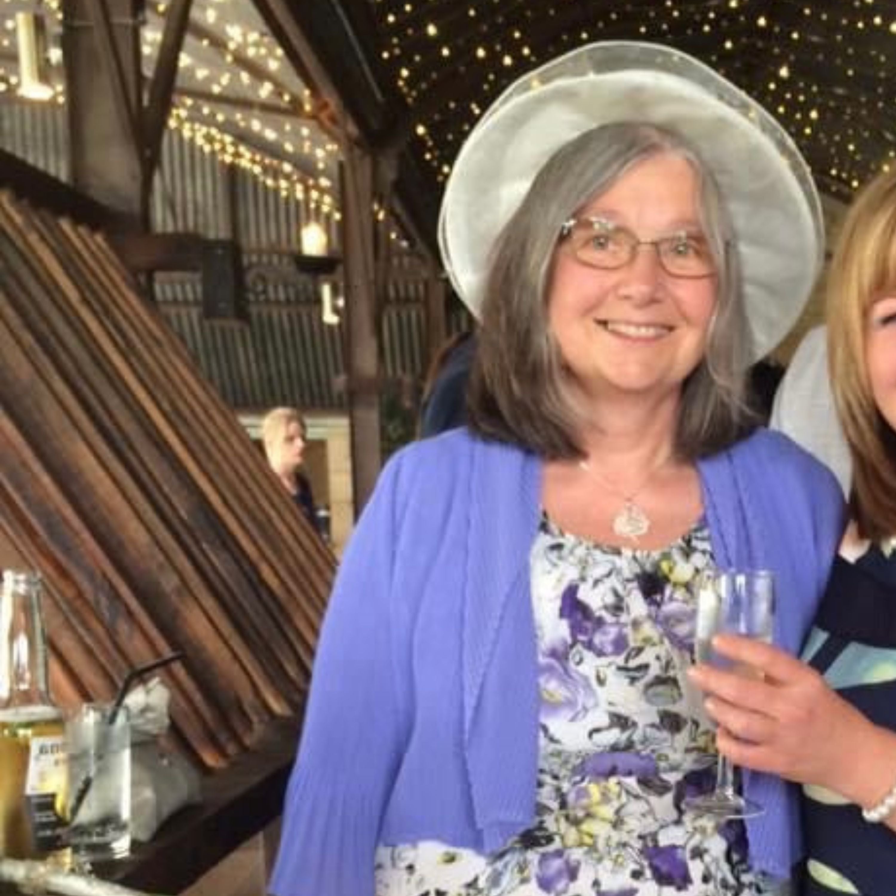 An older woman with grey hair, glasses, and a white hat is smiling. She is wearing a purple cardigan over a white floral dress, standing in a rustic indoor venue with string lights and wooden beams.